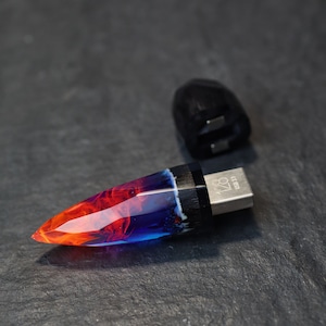 Puede incluir: Una unidad USB de resina azul y naranja con un acento de madera negra y una tapa de metal plateado. La unidad tiene forma de punta y tiene el texto "USB 128" en la tapa de metal.