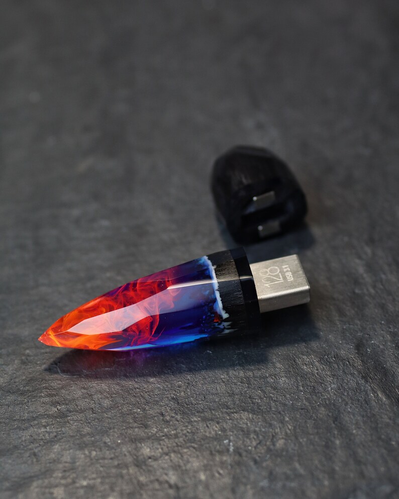 Puede incluir: Una unidad USB de resina azul y naranja con un acento de madera negra y una tapa de metal plateado. La unidad tiene forma de punta y tiene el texto "USB 128" en la tapa de metal.