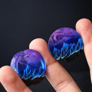 May include: Two round resin cabochons with a blue and purple mountain landscape design. The cabochons are held in a person's hand.