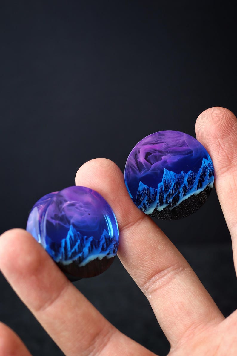 May include: Two round resin cabochons with a blue and purple mountain landscape design. The cabochons are held in a person's hand.