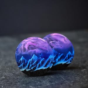 May include: Two round resin plugs with a blue and purple mountain landscape design. The plugs are on a dark gray surface.