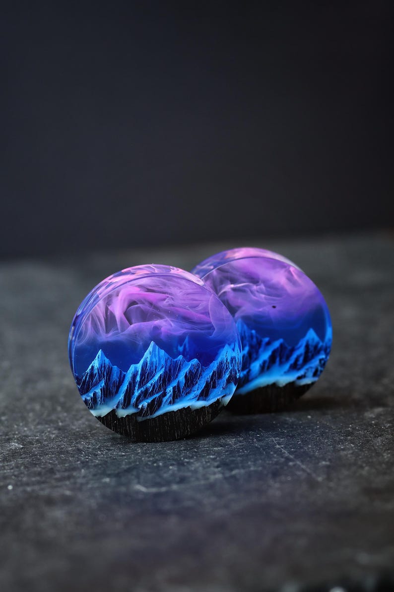May include: Two round resin plugs with a blue and purple mountain landscape design. The plugs are on a dark gray surface.