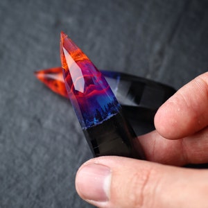 Puede incluir: Un colgante de resina transparente puntiagudo con un patr&oacute;n de remolino de naranja, rojo, azul y negro. El colgante tiene una base oscura, casi negra, con un patr&oacute;n de remolino de resina naranja, roja y azul.