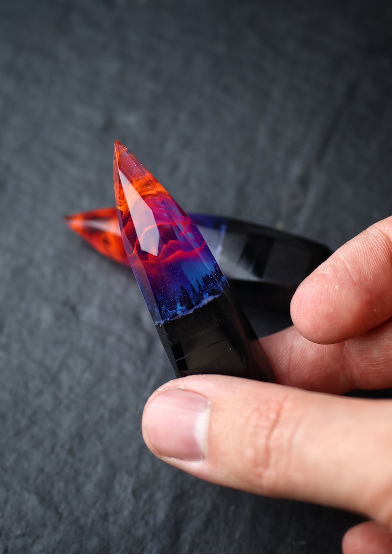 Puede incluir: Un colgante de resina transparente puntiagudo con un patr&oacute;n de remolino de naranja, rojo, azul y negro. El colgante tiene una base oscura, casi negra, con un patr&oacute;n de remolino de resina naranja, roja y azul.