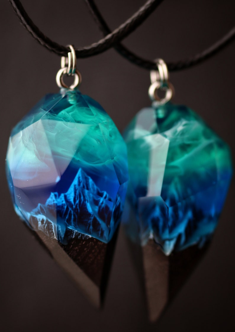 May include: Two pendants featuring a mountain scene in blue and teal resin. The faceted, gem-like pendants have a dark wood base and hang from black cords. The resin art depicts a mountain range, creating a unique jewellery piece.