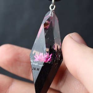 Op de afbeelding: Een heldere, geometrische hangerketting met een zwart koord. De hanger heeft een roze lotusbloem ingekapseld in heldere hars. De onderkant van de hanger is zwart, met de letters "JKM". De hanger wordt door een hand vastgehouden.