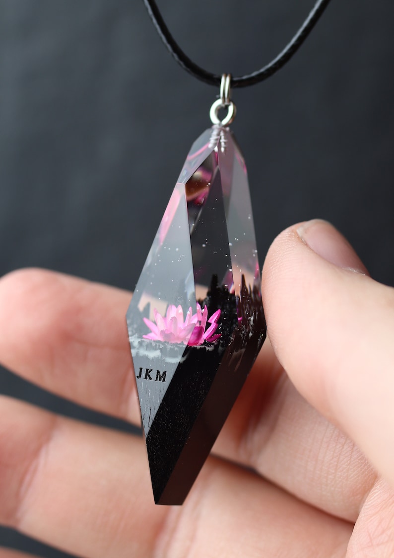 Op de afbeelding: Een heldere, geometrische hangerketting met een zwart koord. De hanger heeft een roze lotusbloem ingekapseld in heldere hars. De onderkant van de hanger is zwart, met de letters "JKM". De hanger wordt door een hand vastgehouden.