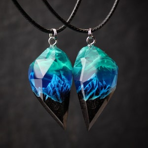 May include: Two matching pendant necklaces. Each pendant features a geometric shape with a dark wood base and a translucent top with blue and teal hues, resembling a mountain landscape. The pendants hang from black cords.