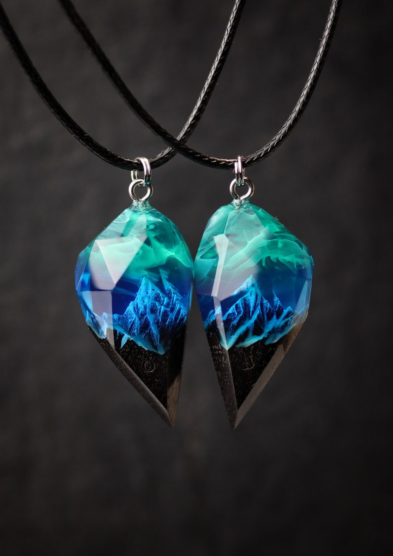 May include: Two matching pendant necklaces. Each pendant features a geometric shape with a dark wood base and a translucent top with blue and teal hues, resembling a mountain landscape. The pendants hang from black cords.