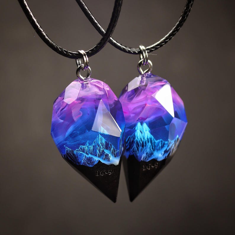 May include: Two matching pendant necklaces. Each pendant has a faceted, heart-shaped design with a black base and a clear, colourful top. The top features a blue and purple landscape with mountain details. The pendants hang from black cords.