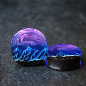 May include: A pair of blue and purple resin ear gauges with a mountain and sky design. The gauges are made of wood and resin.