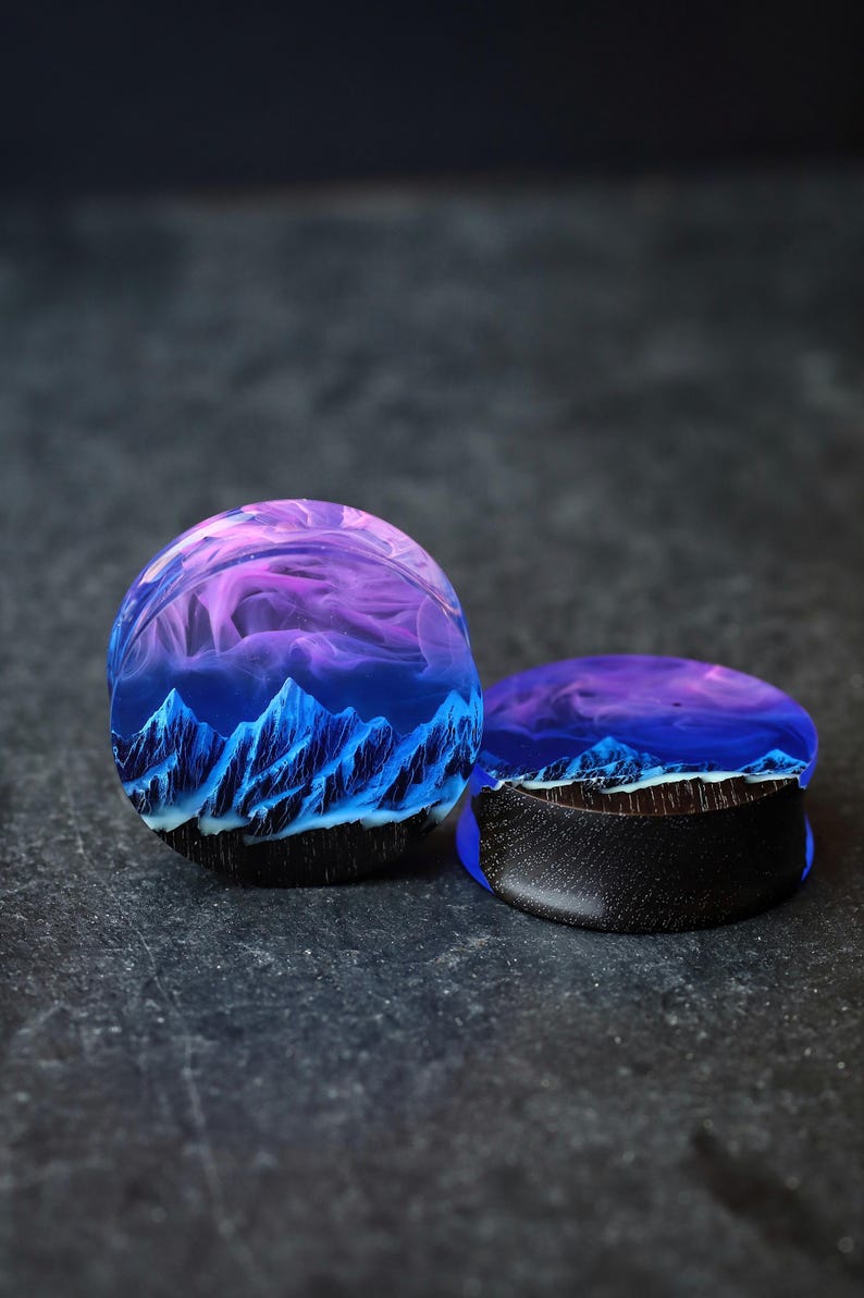 May include: A pair of blue and purple resin ear gauges with a mountain and sky design. The gauges are made of wood and resin.