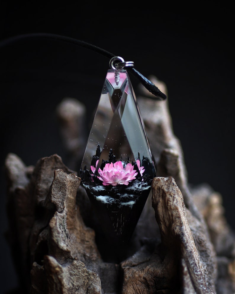 Op de afbeelding: Een heldere, geometrische hangerketting met een roze bloem. De hanger hangt aan een zwart koord. De bloem is geplaatst in een zwarte basis, alles tegen een donkere achtergrond. De hanger is ongeveer 5 cm lang.