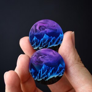 May include: Two round resin earrings with a blue and purple mountain scene inside. The mountains are outlined in black and white, and the sky is a gradient of purple and blue.