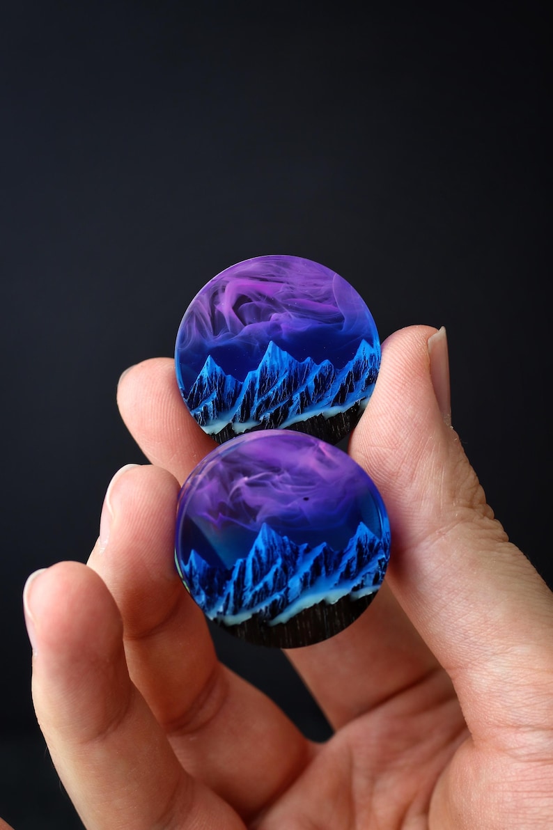 May include: Two round resin earrings with a blue and purple mountain scene inside. The mountains are outlined in black and white, and the sky is a gradient of purple and blue.