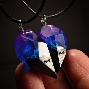 May include: Two matching heart-shaped pendants, each with a faceted, translucent blue and purple resin design. The pendants are connected by black cords and feature the text "16-9" on a black and white surface. The pendants are designed to fit together.