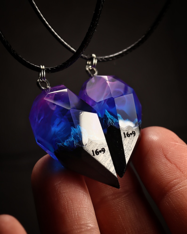 May include: Two matching heart-shaped pendants, each with a faceted, translucent blue and purple resin design. The pendants are connected by black cords and feature the text "16-9" on a black and white surface. The pendants are designed to fit together.