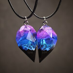 May include: Two matching pendant necklaces. Each pendant has a geometric shape with a black base and a translucent top with purple and blue hues. The top section has a mountain range design. The pendants hang from black cords.