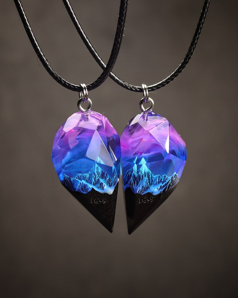 May include: Two matching pendant necklaces. Each pendant has a geometric shape with a black base and a translucent top with purple and blue hues. The top section has a mountain range design. The pendants hang from black cords.