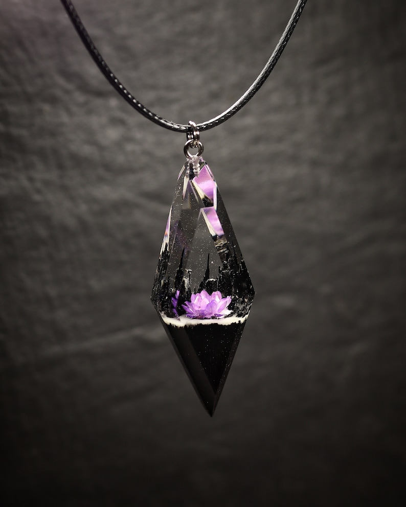 May include: A black cord necklace with a clear resin pendant shaped like a teardrop. Inside the pendant is a purple flower and a black cityscape.