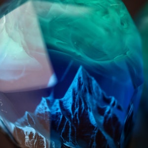 May include: A close-up of a clear, faceted resin object with a mountain range design. The resin is a gradient of blue and teal, with the mountain range in a darker blue. The object has a dark brown base.
