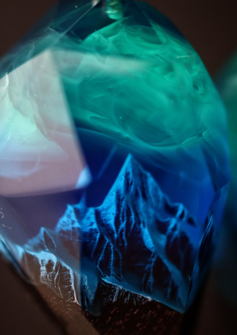 May include: A close-up of a clear, faceted resin object with a mountain range design. The resin is a gradient of blue and teal, with the mountain range in a darker blue. The object has a dark brown base.