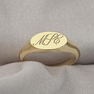 Pode incluir: Um anel de sinete dourado com uma face oval. O anel apresenta iniciais gravadas em uma fonte cursiva. O anel está sobre um fundo de tecido neutro. Uma peça de joalheria clássica.