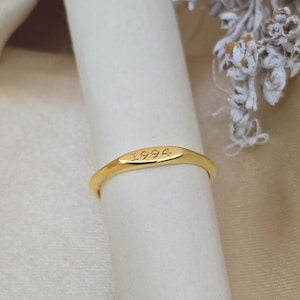 May include: A gold ring with the engraved year "1994". The ring is displayed on a white surface, with a soft, neutral-toned background. The ring has a smooth, polished finish.