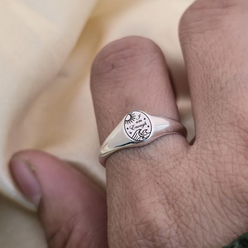 May include: A silver signet ring with a rounded face. The face features an engraved design of a sun, waves, and the words "I am Enough". The ring is worn on a finger.