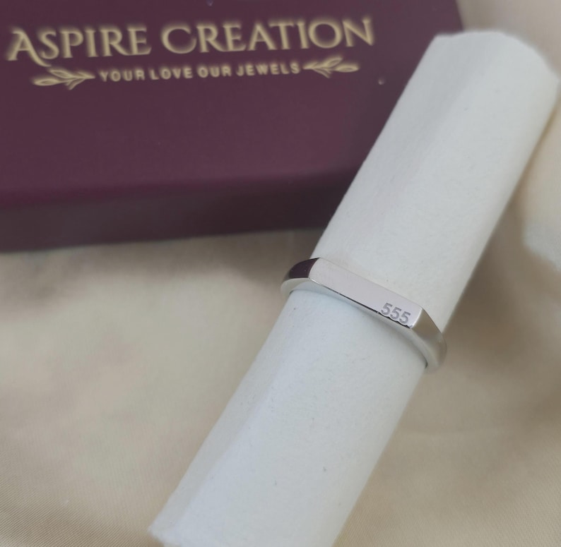 Puede incluir: Un anillo plateado con el n&uacute;mero "555" grabado en la parte delantera. El anillo es rectangular y est&aacute; sobre una superficie blanca. El fondo incluye una caja granate con el texto "ASPIRE CREATION" y "YOUR LOVE OUR JEWELS".