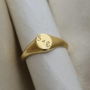May include: A gold signet ring with a smooth, polished finish. The ring features a circular face engraved with the letters "S" and "E", separated by a small heart symbol. The ring is displayed on a white surface.