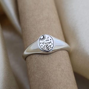 May include: A silver signet ring with an oval face. The face features a sun, waves, and the words "I am Enough" in a black, stylized font. The ring has a smooth, polished finish.