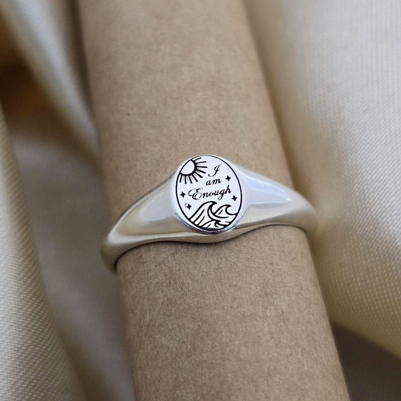 May include: A silver signet ring with an oval face. The face features a sun, waves, and the words "I am Enough" in a black, stylized font. The ring has a smooth, polished finish.
