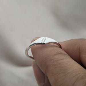 May include: A silver signet ring with a polished finish. The ring features a flat, oval face engraved with the letter "G" in a delicate script. The ring is shown on a finger.