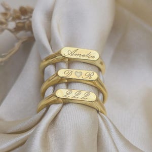以下が含まれることがあります： 刻印された文字が特徴の3つのゴールドカラーのリング。一番上のリングには「Amelia」、真ん中のリングには「D ♡ R」、一番下のリングには「222」と表示されています。リングはクリーム色の生地の上に展示されています。