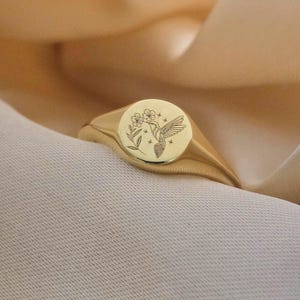 May include: A gold signet ring with a round face featuring an engraved hummingbird and floral design. The ring has a polished finish and a thick band. The ring is displayed on a cream-coloured fabric.