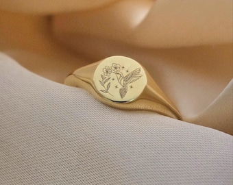 May include: A gold signet ring with a round face featuring an engraved hummingbird and floral design. The ring has a polished finish and a thick band. The ring is displayed on a cream-colored fabric.