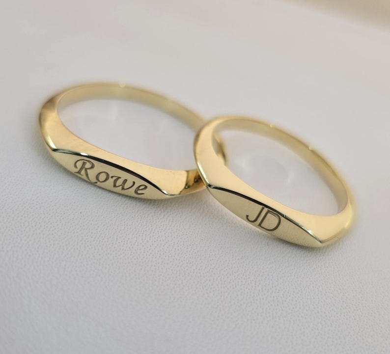 Puede incluir: Dos anillos de oro con texto grabado. Uno tiene grabado "Rowe", el otro "JD". Los anillos son de color oro brillante y tienen un acabado liso y pulido. Los anillos son probablemente para una pareja.