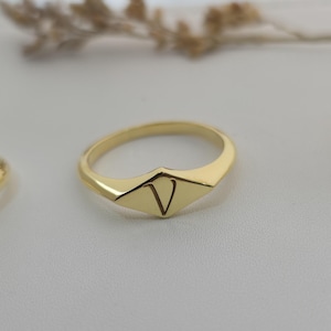 Puede incluir: Un anillo dorado con un dise&ntilde;o geom&eacute;trico. El anillo presenta una cara en forma de diamante con la letra "V" grabada en el centro. El anillo est&aacute; sobre un fondo suave y neutro.