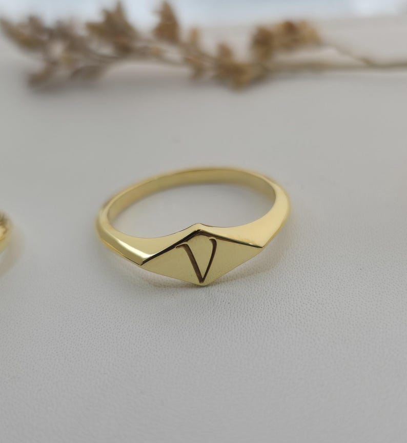 Puede incluir: Un anillo dorado con un dise&ntilde;o geom&eacute;trico. El anillo presenta una cara en forma de diamante con la letra "V" grabada en el centro. El anillo est&aacute; sobre un fondo suave y neutro.