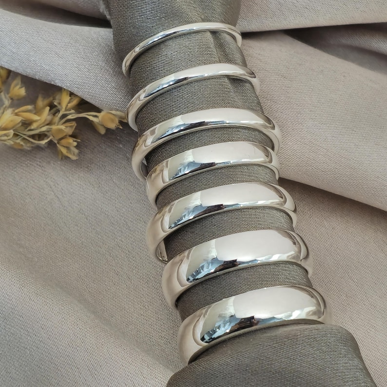 Puede incluir: Un juego de ocho servilleteros plateados y brillantes. Los anillos son lisos y cil&iacute;ndricos, rodeando una servilleta gris. El fondo es una tela gris claro, con unas ramitas de trigo seco.