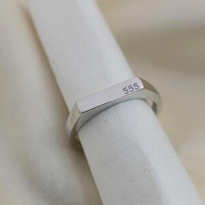 Puede incluir: Un anillo plateado con el n&uacute;mero "555" grabado en una cara rectangular. El anillo tiene un dise&ntilde;o sencillo y moderno con un acabado pulido. El anillo se muestra sobre una superficie blanca.