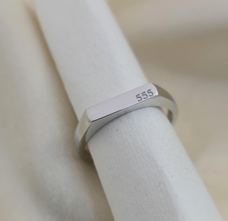 Puede incluir: Un anillo plateado con el n&uacute;mero "555" grabado en una cara rectangular. El anillo tiene un dise&ntilde;o sencillo y moderno con un acabado pulido. El anillo se muestra sobre una superficie blanca.