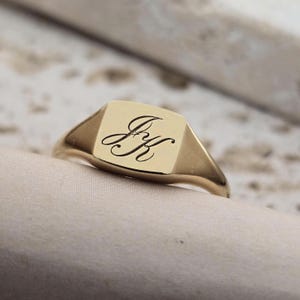 Può includere: Un anello con sigillo d'oro con una faccia quadrata. La faccia dell'anello presenta le iniziali incise "J K" in un carattere corsivo. L'anello è posto su una superficie di colore neutro.