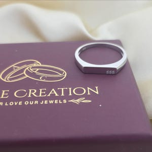 Puede incluir: Un anillo plateado con una cara rectangular, grabado con el n&uacute;mero "555". El anillo est&aacute; sobre una caja morada con texto dorado que dice "IRE CREATION YOUR LOVE OUR JEWELS". El fondo es una tela color crema.