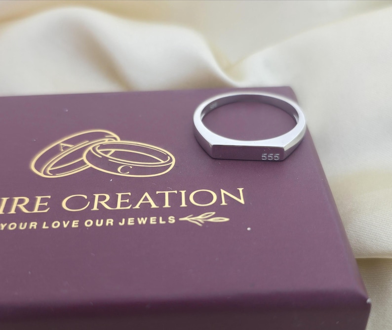 Puede incluir: Un anillo plateado con una cara rectangular, grabado con el n&uacute;mero "555". El anillo est&aacute; sobre una caja morada con texto dorado que dice "IRE CREATION YOUR LOVE OUR JEWELS". El fondo es una tela color crema.