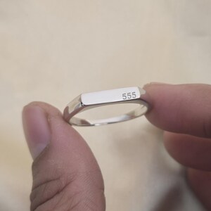 Puede incluir: Un anillo plateado con una parte superior rectangular, grabado con el n&uacute;mero "555". El anillo se sostiene entre dos dedos sobre un fondo neutro. El anillo tiene una superficie pulida y reflectante.