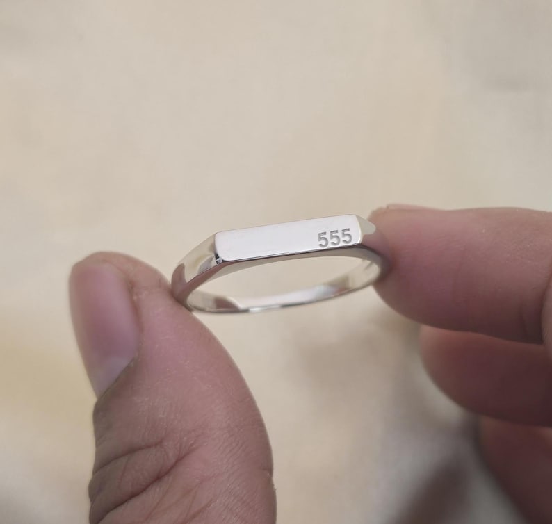 Puede incluir: Un anillo plateado con una parte superior rectangular, grabado con el n&uacute;mero "555". El anillo se sostiene entre dos dedos sobre un fondo neutro. El anillo tiene una superficie pulida y reflectante.