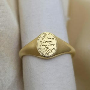 Könnte beinhalten: Ein goldener Siegelring mit ovaler Oberfläche. Die Oberfläche ist mit einer Sonne, Wellen und den Worten "Love Survives Every Storm" graviert. Der Ring wird auf einer weißen Oberfläche präsentiert.