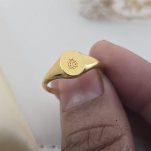 May include: A gold-colored signet ring with a rounded, oval face. The face of the ring features a small, engraved ladybug design. The ring is held by a person's fingers, showcasing its polished finish and simple, elegant design.